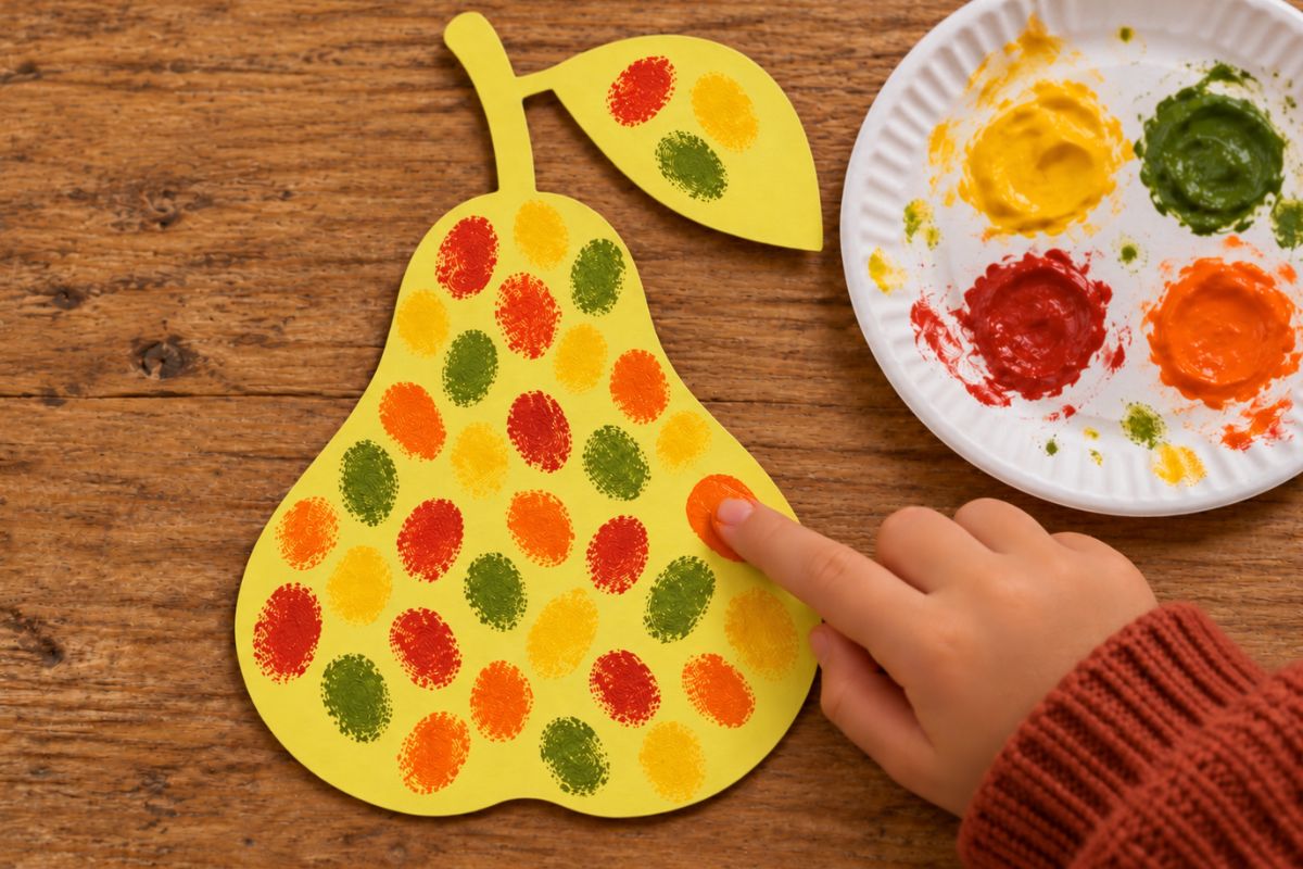 Kind gestaltet beim Herbstbasteln eine Birne aus Bastelpappe mit Fingerabdrücken; die Fingerfarben stehen auf einem Pappteller daneben.