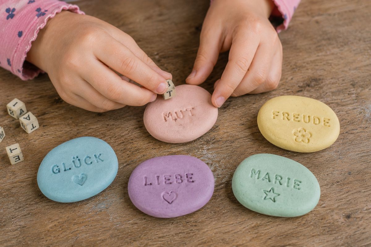 Kind stempelt Wörter in bunte Glückssteine aus Fimo Knete – einfache Geschenkidee mit Mut, Glück, Liebe und Namen