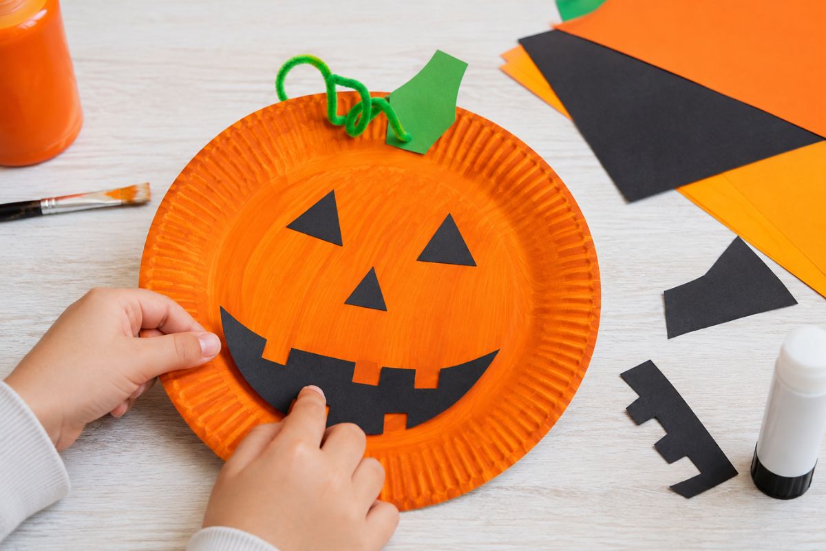 Kinderhände gestalten einen Halloween-Kürbis aus einem orange bemalten Pappteller mit Augen, Nase und Mund aus schwarzem Papier.