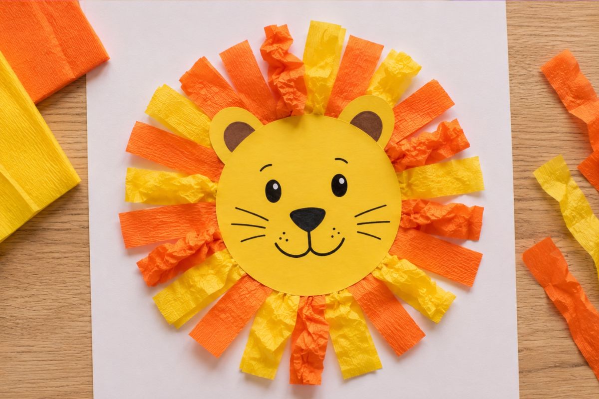 Löwe basteln mit Krepppapier: Von einem Kind gestalteter Löwe mit gelbem Tonpapiergesicht sowie orangefarbenen und gelben Krepppapierstreifen als bunte Mähne