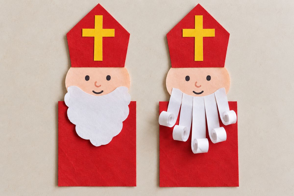 Zwei Nikoläuse aus Papier mit roten Bischofsmützen und gelben Kreuzen, einer mit ausgeschnittenem Bart und einer mit eingerollten weißen Papierstreifen.