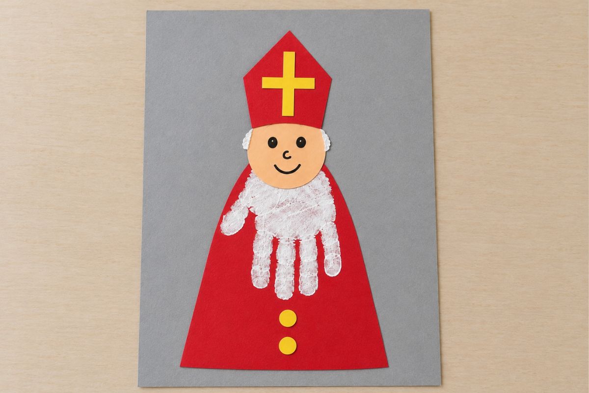 Gebastelter Nikolaus auf grauem Tonpapier mit rotem Papiermantel, weißem Handabdruck als Bart und Bischofsmütze mit gelbem Kreuz.