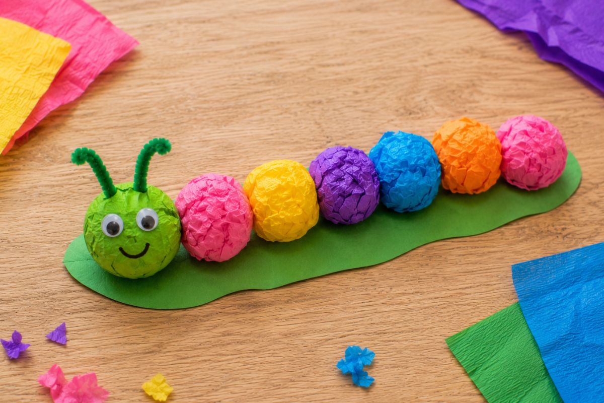 Raupe basteln mit Krepppapier: Von einem Kind gebastelte Raupe aus bunten geknüllten Krepppapierkugeln mit Wackelaugen und Pfeifenreiniger-Fühlern auf grünem Tonpapier