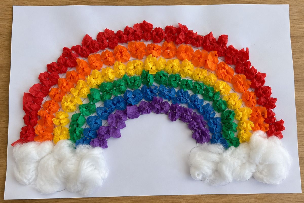 Regenbogen basteln mit Krepppapier: Kind klebt geknüllte Krepppapierstücke auf einen Regenbogen aus Tonpapier.