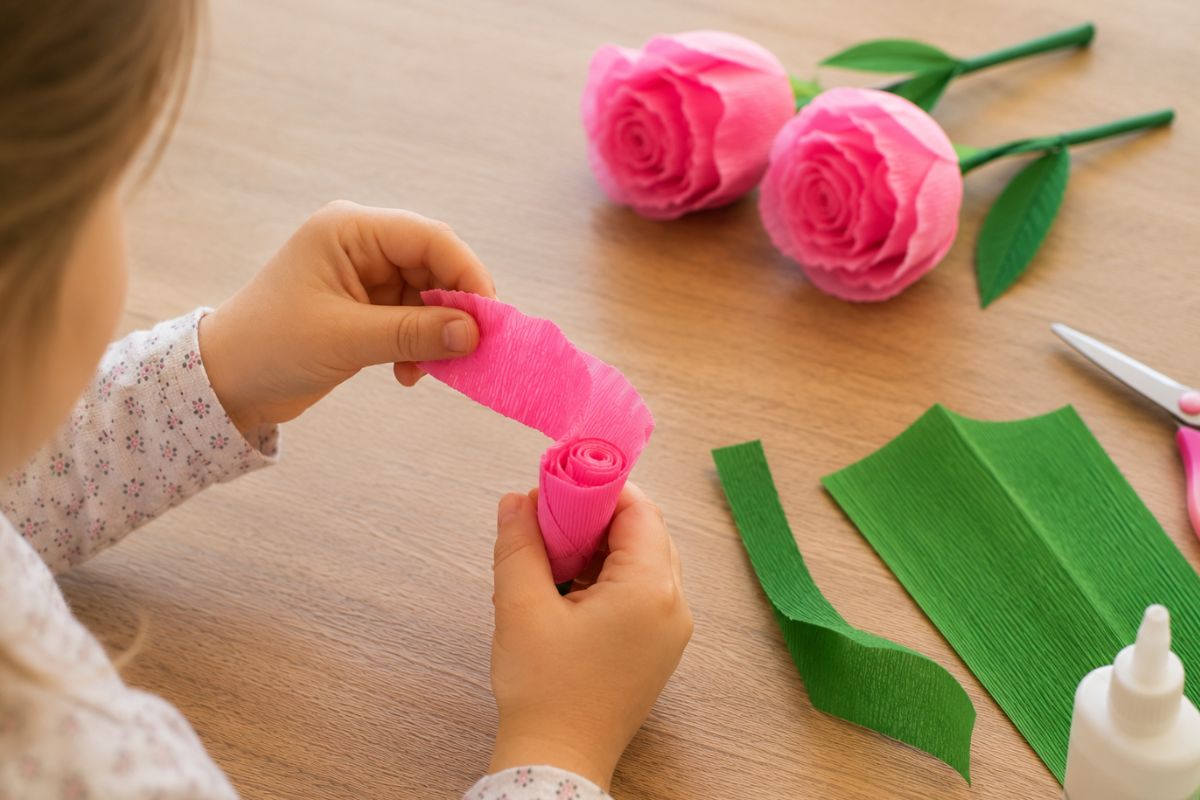 Rosen basteln mit Krepppapier: Kind rollt einen gedehnten Krepppapierstreifen zu einer Rose auf, im Hintergrund liegen fertige Krepppapierrosen.