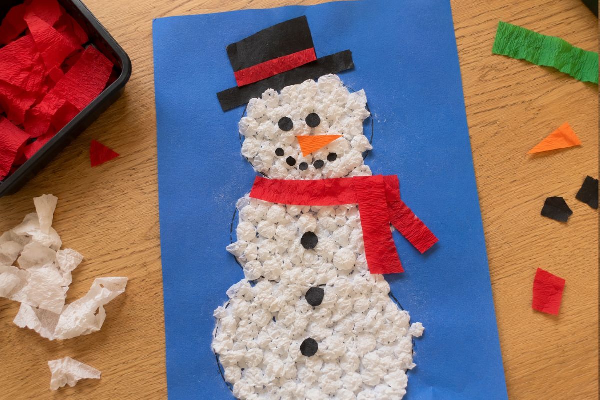 Schneemann basteln mit Krepppapier: Von einem Kind gestalteter Schneemann mit locker und unregelmäßig geknüllten weißen Krepppapierkugeln sowie Hut, Schal, Nase und Knöpfen aus Tonpapier